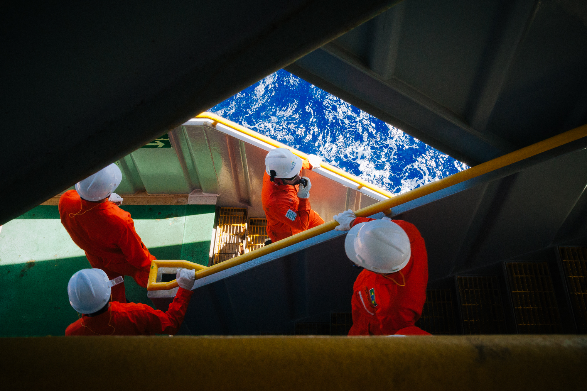 Colaboradores vestidos com macacões laranjas e capacetes brancos, descendo escadas em plataforma